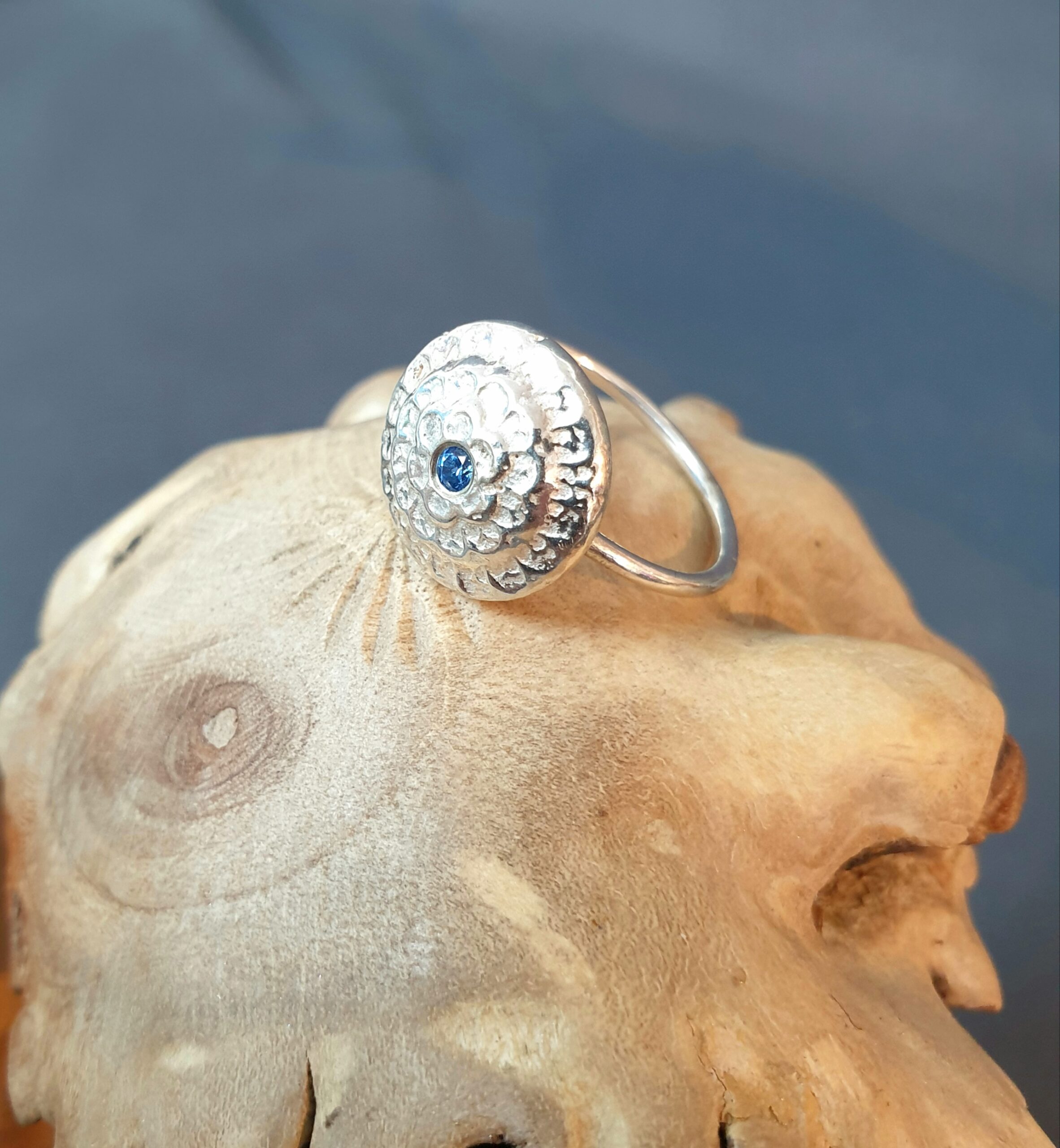 This image showcases a handcrafted silver ring with a circular, domed face decorated in an intricate, textured pattern that radiates outward in a floral-like arrangement, drawing the eye to a small, deep blue gemstone set at the centre; the softly polished surface highlights subtle variations in tone and texture, emphasising the artisanal nature of the piece, while the slim, simple band provides a clean contrast to the detailed top; the ring is carefully positioned on a natural, sculptural wooden surface with visible grain and organic contours, adding warmth and an earthy character, while the blurred, neutral background enhances the sense of depth and keeps the focus on the ring’s distinctive design, refined craftsmanship, and unique handmade appeal.