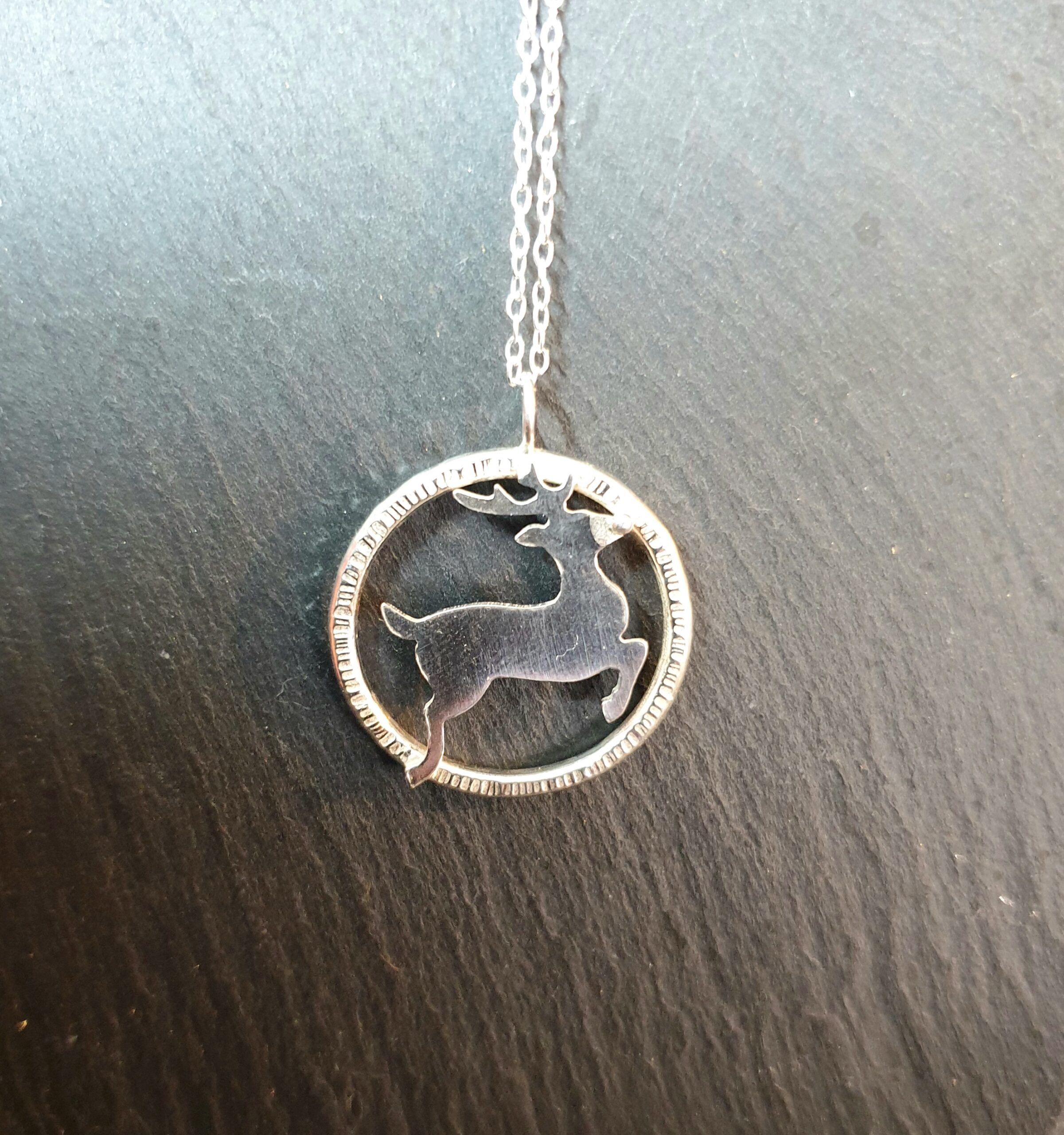 A stylish silver necklace featuring a circular pendant with a finely cut reindeer silhouette is beautifully displayed against a dark, natural surface, creating a striking contrast that enhances its polished finish. The smooth, reflective metal catches the light, highlighting the subtle texture around the pendant’s edge and the clean, graceful lines of the design. The delicate chain extends upward, adding to the piece’s elegant simplicity while keeping the focus on the intricate craftsmanship of the pendant. This refined presentation showcases the necklace as a unique and timeless accessory, perfect for adding a subtle, nature-inspired detail to any outfit or for giving as a thoughtful, handcrafted gift.