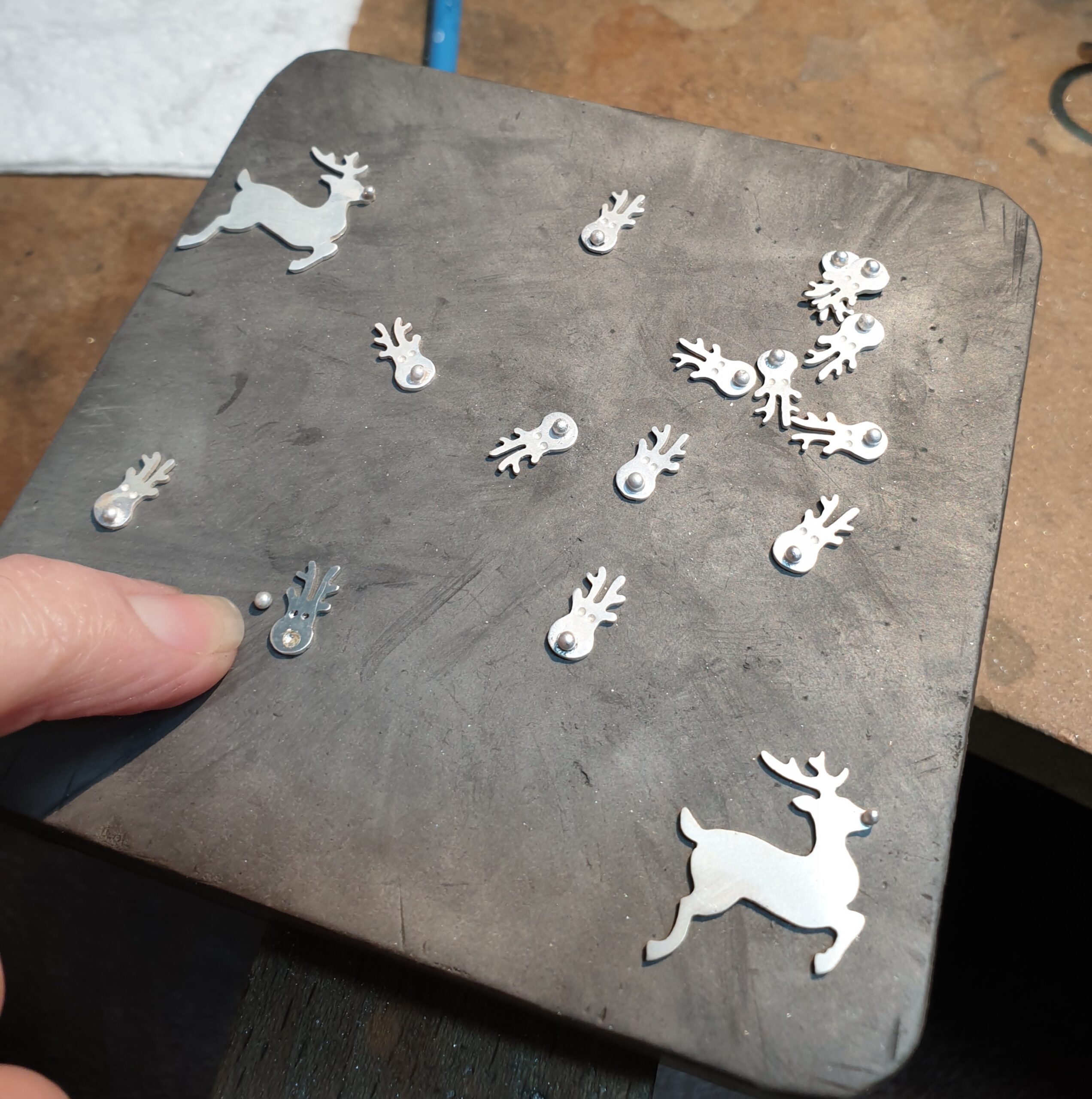 This behind-the-scenes image captures the meticulous craftsmanship involved in jewellery making, showcasing a workbench surface with multiple small, hand-cut metal shapes arranged across a soldering block. Among the pieces are miniature decorative forms and a larger reindeer silhouette, each with carefully placed raised details, highlighting the precision and care taken during fabrication. A fingertip in the foreground provides scale, emphasizing the delicate size of each component, while the textured work surface and scattered tools subtly reveal the authentic workshop setting. The natural lighting and raw materials convey a sense of artistry in progress, offering a glimpse into the skill and attention to detail that goes into creating unique, handcrafted pieces.