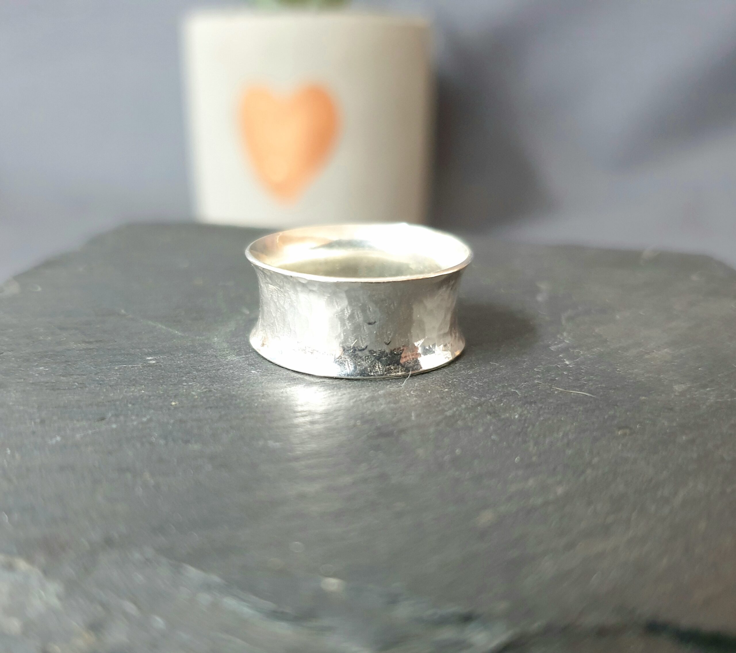 This image highlights a handcrafted silver ring placed on a dark, natural stone surface, with its gently flared silhouette and lightly hammered texture reflecting soft light to reveal subtle variations in tone and finish; the simple composition draws focus to the ring’s artisanal quality, while the softly blurred background, featuring a pale plant pot with a faint heart motif, adds a warm and understated decorative touch, creating a calm, minimalist setting that enhances the ring’s refined yet organic character.
