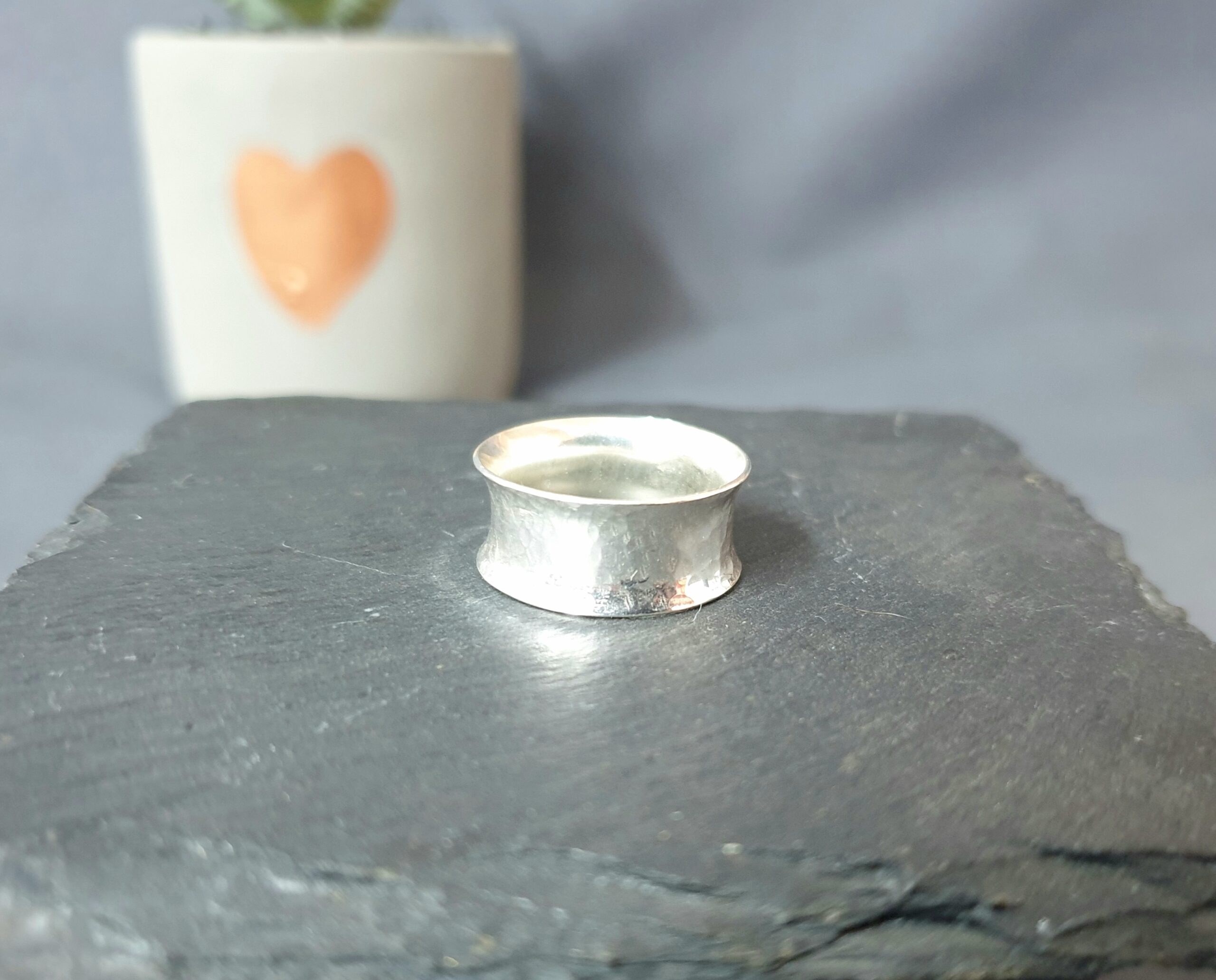 This image features a handcrafted silver ring displayed on a dark, textured stone surface, its gently flared shape and softly hammered finish catching the light to create subtle reflections that emphasise its artisanal character; in the blurred background, a small white plant pot with a simple heart design adds a warm, personal touch while remaining out of focus to keep attention on the ring, and the neutral tones combined with soft lighting create a calm, minimalist aesthetic that highlights the ring’s simplicity, elegance, and unique handmade quality.