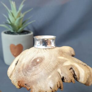 This image presents a close-up of a handcrafted silver ring resting atop an organic, sculptural wooden surface with natural knots, cracks, and a softly carved, petal-like edge, highlighting the contrast between the smooth, reflective metal and the textured warmth of the wood; in the softly blurred background, a small potted succulent with pale green leaves sits in a light-colored pot decorated with a simple heart motif, adding a subtle touch of homeliness and depth, while the neutral grey backdrop enhances the clean, modern aesthetic and draws full attention to the ring as the central focal point.