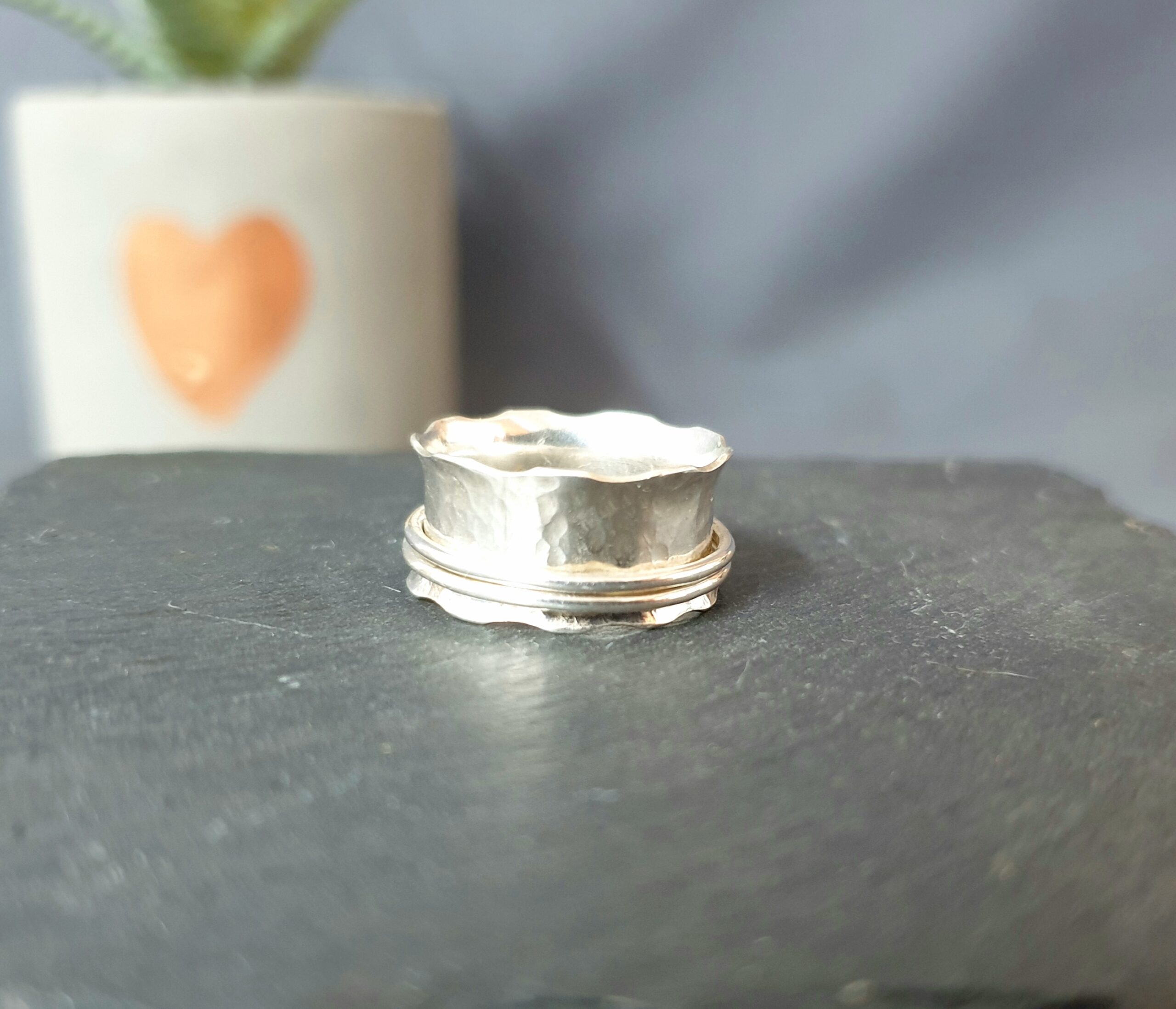 This image features a handcrafted silver ring displayed on a smooth, dark surface, beautifully highlighting its organic design and artisanal finish. The ring showcases a softly hammered texture with a gently scalloped edge, creating a fluid, irregular silhouette that reflects its handmade nature. A subtle double-band detail encircles the base, adding depth and a refined layered element while maintaining a clean, modern look. The polished silver surface catches the light, enhancing the texture and contours, while the softly blurred background—featuring a muted plant and a warm-toned decorative accent—adds a calm, natural setting that draws attention to the craftsmanship and elegance of this unique, versatile piece.
