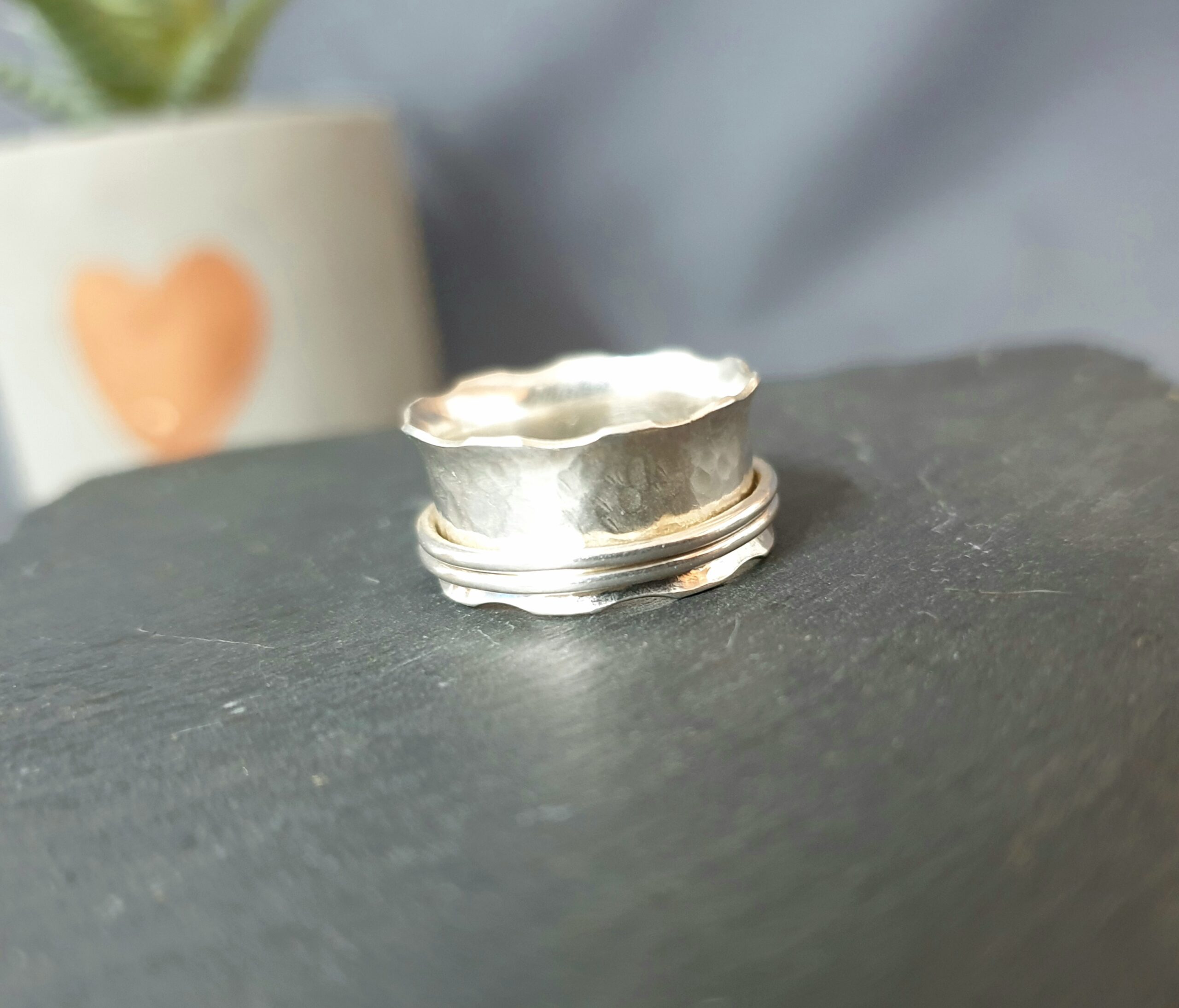 This image showcases a handcrafted silver ring displayed on a smooth, dark surface, emphasising its minimalist yet distinctive design. The ring features a softly hammered texture paired with a gently scalloped edge, creating an organic, slightly irregular silhouette that highlights its handmade quality. A subtle double-band detail wraps around the base, adding depth and structure while maintaining a clean, modern aesthetic. The polished silver finish catches and reflects light beautifully, enhancing the texture and form, while the softly blurred background—featuring muted tones and a hint of natural décor—keeps the focus on the ring, making it an elegant and versatile piece perfect for everyday wear or as a unique, thoughtful gift.