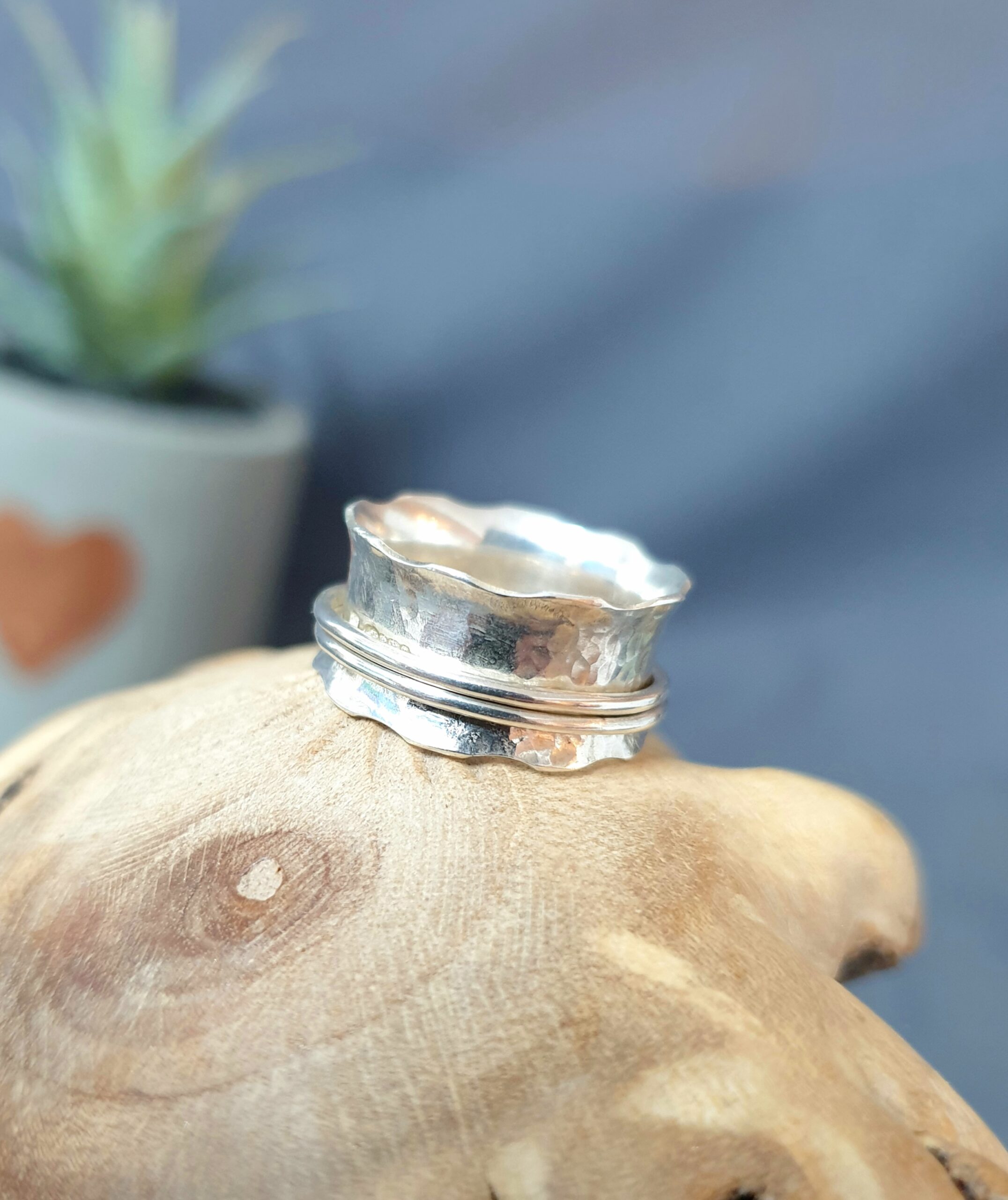 This image presents a handcrafted silver ring displayed on a natural wooden surface, highlighting its organic, artisanal design and subtle craftsmanship. The ring features a softly hammered texture with a gently scalloped edge that creates an uneven, flowing silhouette, giving the piece a unique and handmade character. A delicate double-band detail wraps around the base, adding structure and visual interest while complementing the ring’s rustic yet refined aesthetic. The polished silver surface reflects soft light, enhancing the texture and contours, while the blurred background keeps the focus on the ring itself, making it an elegant and distinctive accessory perfect for everyday wear or a meaningful gift.