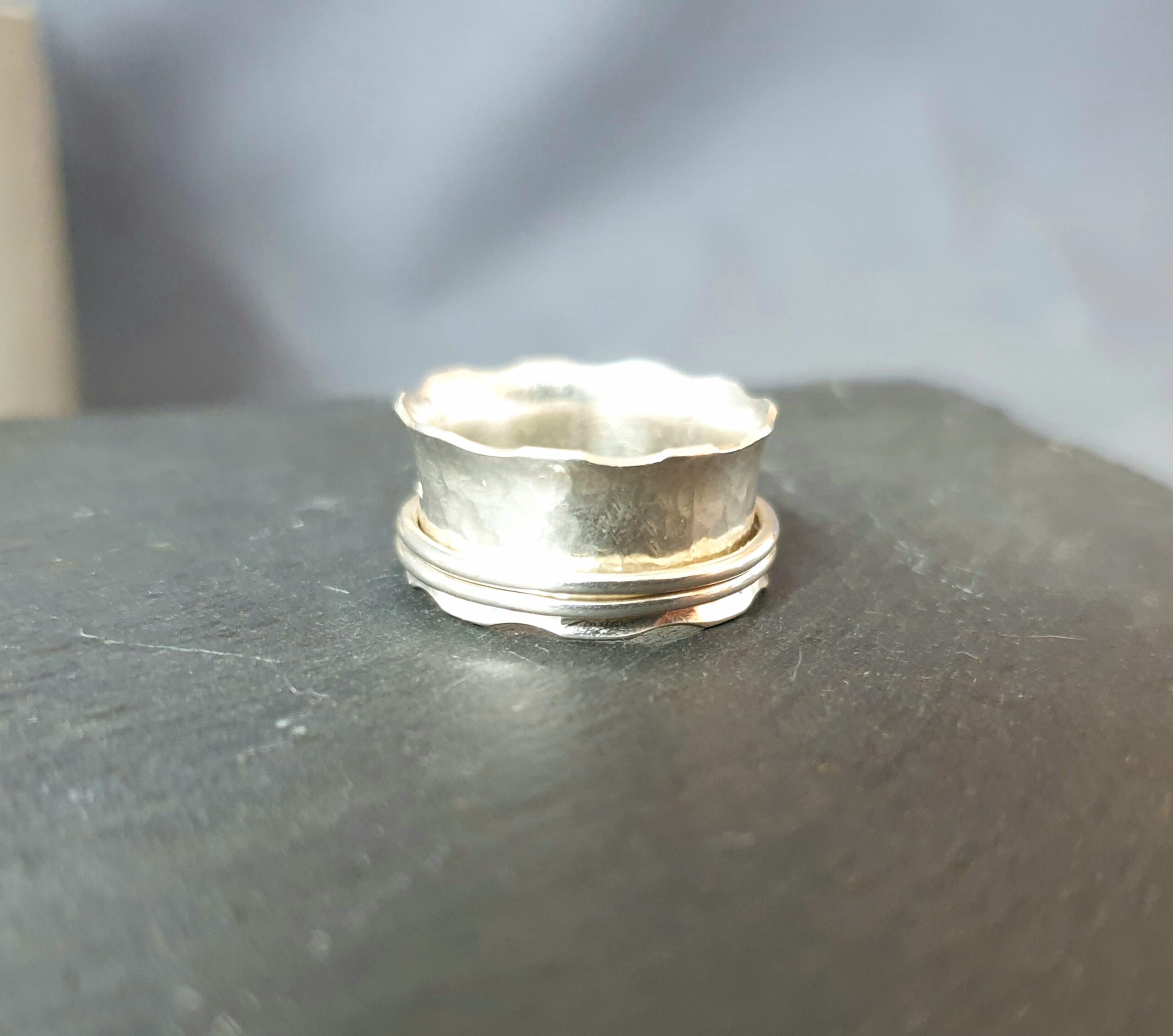 This image showcases a handcrafted silver ring displayed on a dark surface, highlighting its softly hammered texture and distinctive scalloped edge that gives the piece a natural, organic feel. The ring features a subtle double-band detail at the base, adding depth and structure to the design while maintaining a minimalist aesthetic. Its polished finish reflects the light beautifully, emphasising the unique character and artisanal craftsmanship of the piece. The softly blurred background keeps the focus on the ring’s intricate detailing, making it an elegant and versatile accessory that pairs timeless simplicity with a modern, handcrafted edge.