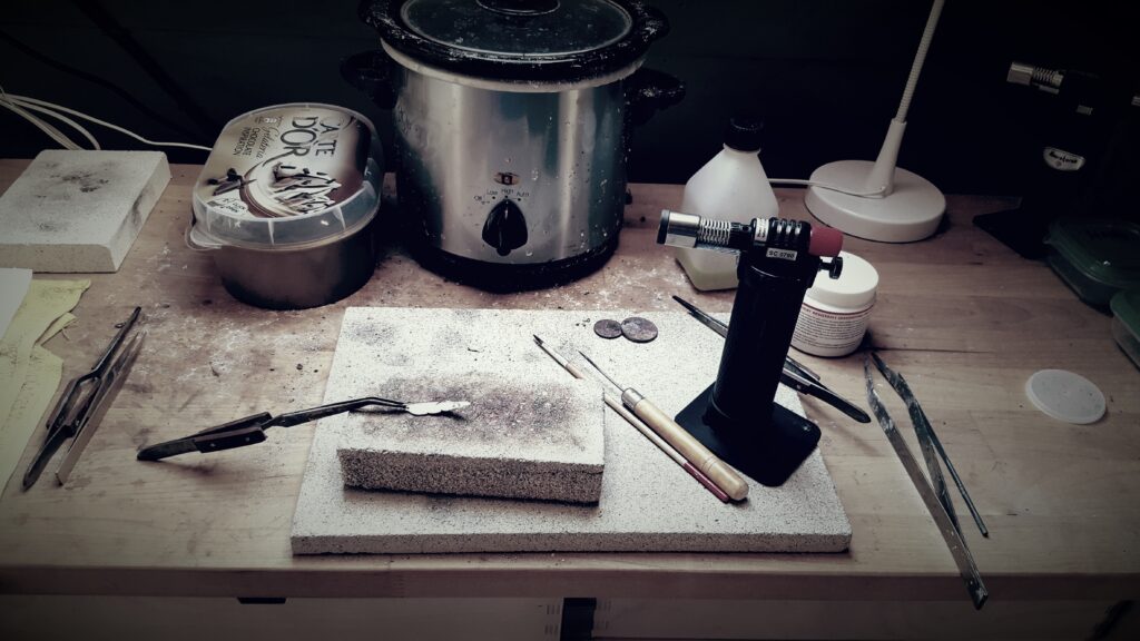 This image shows a jeweller’s workbench prepared for detailed metalworking and jewellery creation. At the centre, two stacked heat-resistant blocks display dark scorch marks and residue from repeated soldering work, with a pair of tweezers holding a small silver component in place. To the right, a compact blowtorch stands ready for precision heating, surrounded by fine tools such as files, picks, and a slim-handled instrument used for delicate adjustments.